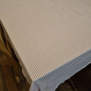 May include: A rectangular tablecloth featuring a striped pattern in white and light brown. The fabric is draped over a wooden table, with a corner of the cloth visible.