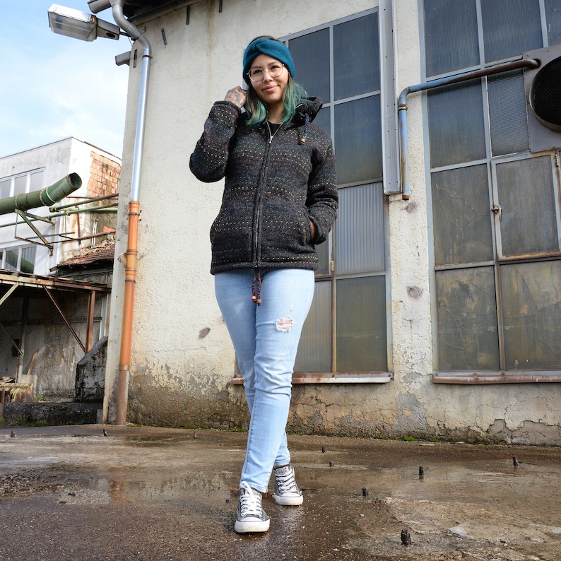 May include: A woman wearing a dark gray and black patterned jacket with a teal blue beanie and light blue jeans. She is standing in front of a building with large windows.