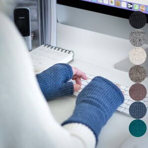 Könnte beinhalten: Eine Person trägt blaue fingerlose Handschuhe, während sie auf einer weißen Tastatur tippt. Die Handschuhe sind aus weichem, gestricktem Stoff. Die Tastatur befindet sich auf einem weißen Schreibtisch. Das Bild zeigt mehrere Farbmuster der Handschuhe.
