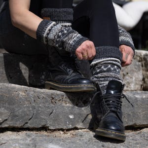 Könnte beinhalten: Ein Paar dunkelgraue Strickstulpen mit einem weißen und hellgrauen Fair-Isle-Muster. Die Stulpen werden über einer schwarzen Leggings getragen und mit schwarzen Schnürstiefeln aus Leder kombiniert. Der Strickstoff hat ein geripptes Bündchen.
