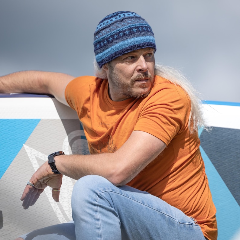 May include: A man wearing a blue and grey patterned beanie, an orange t-shirt, and blue jeans sits on a blue and white surface.