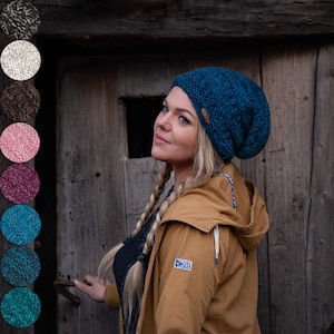 Könnte beinhalten: Eine Frau mit langen blonden Haaren trägt eine blaue Strickmütze und eine braune Jacke. Sie steht vor einer Holztür.