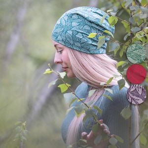 Könnte beinhalten: Eine Frau trägt eine türkisfarbene Mütze mit einem weißen Blatt-Muster. Die Mütze ist aus einem weichen, dehnbaren Stoff. Die Frau steht in einer Waldumgebung.