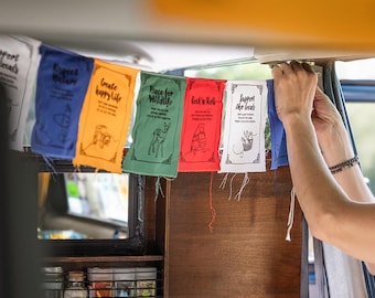 May include: A string of colorful fabric prayer flags with positive messages, including "Support Local", "Create Happy Life", "Peace for Wildlife", "Rock n Roll", and "Support the Locals". The flags are hanging from the ceiling of a vehicle.