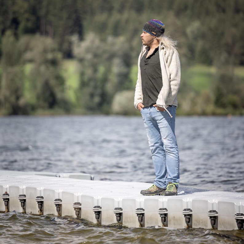May include: A man wearing a white jacket, a black shirt, blue jeans, and a colorful knit beanie stands on a floating dock. The dock is made of white plastic and has multiple black metal clips.