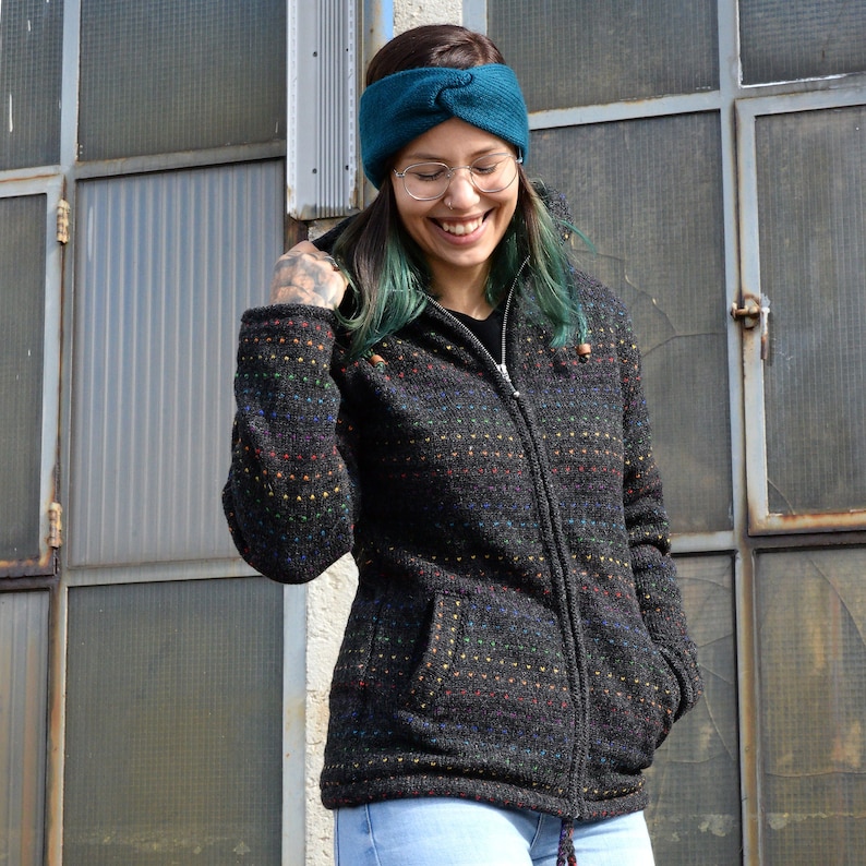 May include: A woman wearing a black hooded jacket with colorful stripes and a teal headband. She is smiling and looking down.