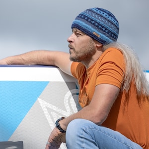 May include: A man wearing a blue and grey patterned beanie sits on a blue and white patterned inflatable paddle board. He is wearing a bright orange t-shirt and blue jeans.