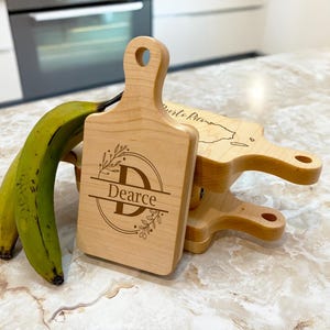 May include: Wooden cutting boards with handles, stacked on a countertop. The front board is engraved with the name "Dearce" and a "D" initial. Two green bananas are on the left. The boards are light brown.