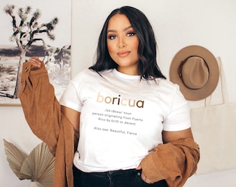 May include: White t-shirt with the word "boricua" in gold lettering, along with a definition. The shirt also includes the phrases "person originating from Puerto Rico by birth or decent" and "Also see: Beautiful, Fierce."