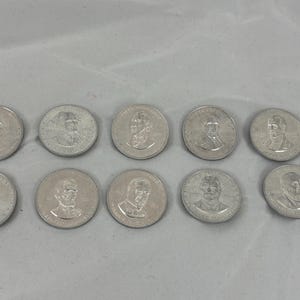May include: Ten silver coins with portraits of historical figures. The coins are round and have a detailed relief design. The coins are arranged on a light gray surface.