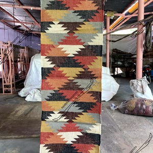 May include: A long, narrow rug with a geometric pattern in shades of red, orange, yellow, brown, black, and gray. The rug has a fringe on both ends.