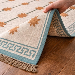 May include: Beige and blue area rug with a starburst pattern. The rug has a blue border with a Greek key design and fringe. The rug is made of a woven material and is displayed on a hardwood floor.