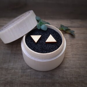 Puede incluir: Un par de pendientes triangulares de madera en una pequeña caja redonda de madera con interior de terciopelo negro. Los pendientes son de color marrón claro.