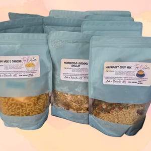 May include: Several light blue resealable pouches filled with food mixes. The pouches have clear windows showing the contents, including pasta and vegetables. Each pouch has a label with text, including "Creamy Mac & Cheese," "Homestyle Lasagna Skillet," and "Alphabet Soup Mix."