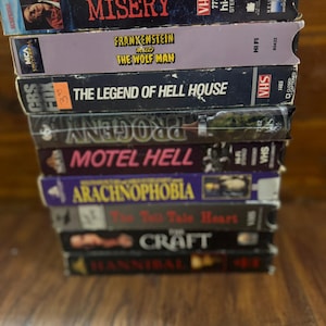 May include: A stack of VHS tapes featuring horror and thriller movie titles. Titles include "Misery", "Frankenstein Meets The Wolf Man", "The Legend of Hell House", "Motel Hell", "Arachnophobia", "The Tell Tale Heart", "Craft", and "Hannibal".