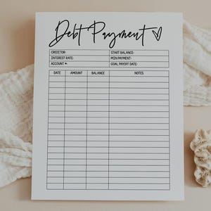 May include: A white paper with the words "Debt Payment" in cursive script. The paper has sections for creditor, interest rate, account, date, amount, balance, notes, start balance, min payment, and goal payoff date. A heart symbol is next to the title.
