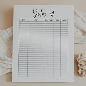 May include: A white sales tracker sheet with the word "Sales" in cursive script and a heart symbol. The sheet has columns for date, item, sale price, fees, and profit. A white cloth and a piece of coral are in the background.