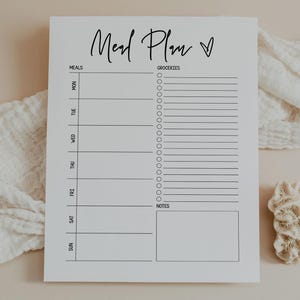 Może przedstawiać: Biały notatnik z planem posiłków ze słowami "Meal Plan" w kursywie. Notatnik ma sekcje na posiłki, artykuły spożywcze i notatki. Dekoracyjny koral i biała tkanina znajdują się w tle.