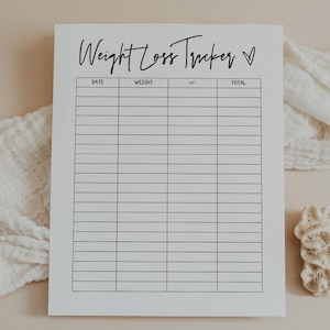 Puede incluir: Un rastreador de pérdida de peso blanco con el título escrito a mano "Weight Loss Tracker" y un símbolo de corazón. El rastreador tiene columnas para fecha, peso, +/- y total. Un paño blanco y un trozo de coral están en el fondo beige.
