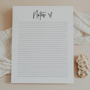 Può includere: Un blocco note bianco con linee orizzontali e la parola "Notes" in un carattere scritto a mano, con un simbolo a forma di cuore. Il blocco note è su una superficie dai toni neutri, con un panno bianco e un pezzo di corallo nelle vicinanze.