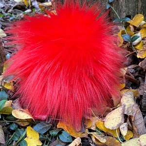 May include: A bright red fluffy pom pom, likely made of faux fur, is lying on a bed of fallen leaves. The pom pom is a vibrant red color and has a soft, fuzzy texture.
