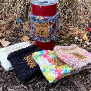May include: Four colorful knitted coffee cup cozies with wooden tags. The tags have different sayings about coffee. One tag says "Sorry What Is Before Coffee", another says "OCD Obsessive Coffee Disorder", and another says "Drop it like it's hot".