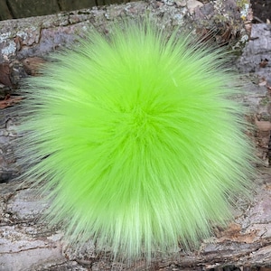 May include: A bright green fluffy pom pom made of faux fur. The pom pom is sitting on a piece of wood.