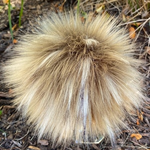 May include: A fluffy, light brown and white faux fur pom pom. The pom pom is made of soft, synthetic fibers and has a round shape.