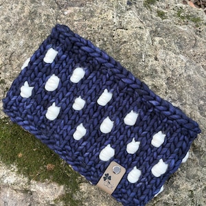 May include: A chunky knit headband in navy blue with white, raised dot accents. The headband features a small, rectangular leather tag with the text "abk" and a four-leaf clover symbol. The item is displayed on a textured stone surface.