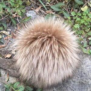May include: A fluffy, light brown faux fur pom pom. The pom pom is round and has a soft, plush texture.