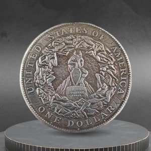 May include: A detailed silver coin with relief artwork, displaying figures and text. The coin is inscribed with "UNITED STATES OF AMERICA" and "ONE DOLLAR". The design includes a central shield and surrounding figures. The coin is presented on a tiered platform.