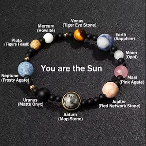 May include: A planet-themed beaded bracelet with stones representing planets like Tiger Eye and Sapphire, strung on a black cord with gold accents. The bracelet features the text "You are the Sun".