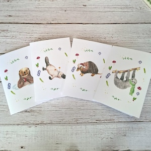 以下が含まれることがあります： 動物の水彩画が描かれた4枚の白いグリーティングカード。マグカップを持つカワウソ、星を持つカモノハシ、サンタ帽をかぶったアルマジロ、緑のマフラーをして枝にぶら下がるナマケモノが描かれており、周囲には植物のモチーフが添えられています。