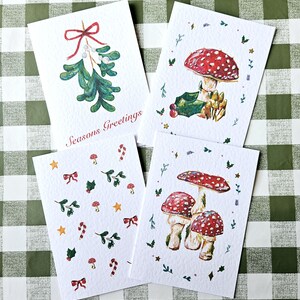 以下が含まれることがあります： お祝いのデザインが施された4枚の水彩グリーティングカード。1枚のカードにはヤドリギと「Seasons Greetings」の文字が描かれています。他のカードには、キノコ、ヒイラギ、リボン、キャンディケインが描かれています。カードは緑と白のチェック柄の表面にあります。