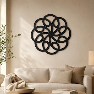 May include: A black, circular wall art piece with an intricate, interwoven design. The artwork is mounted on a neutral-colored wall above a beige sofa with matching pillows. The room has a minimalist, modern aesthetic.