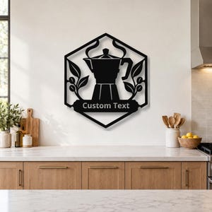 Könnte beinhalten: Ein schwarzes Wandkunstobjekt aus Metall in einem sechseckigen Rahmen mit der Silhouette einer Espressokanne, stilisierten Kaffeeblättern und einem Banner am unteren Rand mit dem Text Custom Text. Das Design ist symmetrisch mit Dampf.