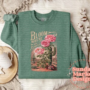 May include: A green long-sleeved shirt featuring the text "Bloom Where You Are Planted" and a vintage illustration of pink flowers and a cactus.