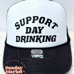 May include: White trucker hat with black mesh back. The front of the hat has black text that reads "SUPPORT DAY DRINKING".
