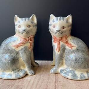 May include: Two ceramic cat figurines featuring a cream-coloured base with grey tabby-style markings and painted orange bows around their necks. Each cat is depicted in a seated position with pointed ears, whiskers, and orange-toned eyes.