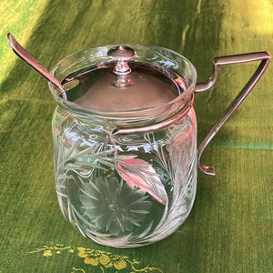 May include: A clear glass sugar bowl with a silver lid and handle. The bowl has an etched floral design and comes with a matching silver spoon.