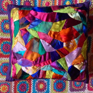 May include: A cushion with a patchwork design. It features irregularly shaped fabric pieces in pink, orange, yellow, green, and purple. The cushion has a dark purple border and is placed on a colourful crocheted blanket.