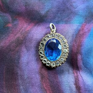 May include: An oval pendant featuring a large, bright blue gemstone encircled by smaller, clear stones. The pendant has a silver-toned metal setting and a small loop for a chain. The backdrop is a colourful, swirled fabric.