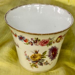 May include: A white porcelain cup with a gold rim and floral pattern. The cup features a variety of flowers in shades of pink, yellow, blue, and purple.