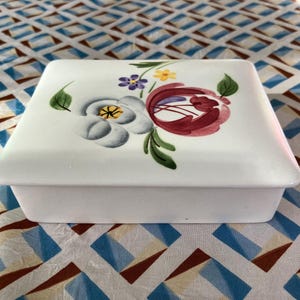 May include: White ceramic box with a floral design. The flowers are painted in shades of red, blue, yellow, and green.