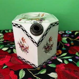 May include: A white, hexagonal ceramic container with floral designs and a black and white checkered border. The container has a dark metal fixture and is decorated with colourful flowers. The background is a green wall and a black floral fabric.