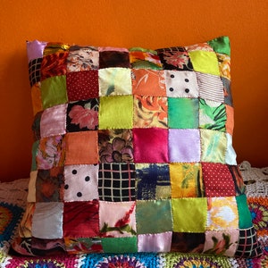 May include: A colourful patchwork cushion featuring a variety of floral and geometric patterns in shades of red, green, pink, yellow, and brown. The cushion is made of satin fabric and has a square shape.