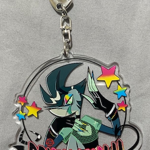 May include: Clear acrylic keychain featuring a stylized character with teal and black hair, wearing a green mesh top. The keychain has the text "PRIDEMONTH" in rainbow colors, surrounded by colorful stars and a silver clasp.