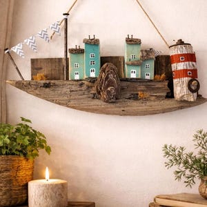 Könnte beinhalten: Rustikale Wanddekoration in Bootsform aus Treibholz, mit Miniaturhäusern in Blau- und Grüntönen, einem Leuchtturm und einer dekorativen Muschel. Eine kleine Flagge und ein Seil ergänzen das nautische Thema. Eine brennende Kerze und Pflanzen sind im Vordergrund.