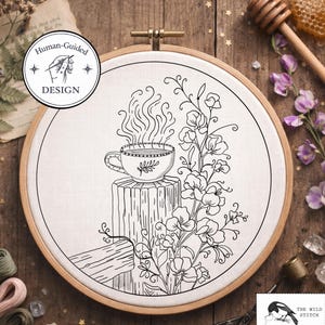 Könnte beinhalten: Ein Stickrahmen mit einem schwarz-weißen Design einer Teetasse auf einem Holzpfosten, aus der Dampf aufsteigt. Das Design enthält florale Elemente und den Text "Human-Guided Design". Das Logo The Wild Stitch ist sichtbar.