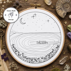 May include: An embroidery hoop featuring a black and white landscape design. The artwork depicts a house, a crescent moon, stars, and a field of flowers. The text "Human-Made Design" is also included.